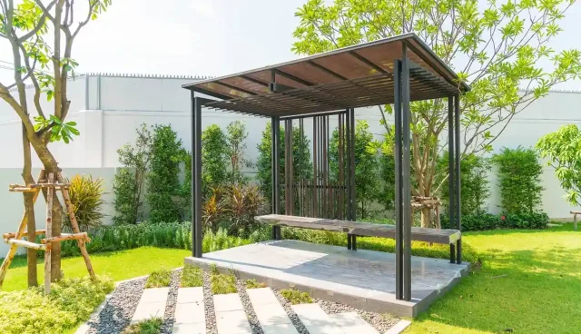 Pergolado com telhas e um banco para sentar em um jardim verde e caminhos de pedra.