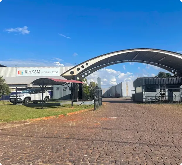 Fachada da empresa Biazam com portão em arco, guarita, carro Jeep branco estacionado e pavimentação de paralelepípedo.